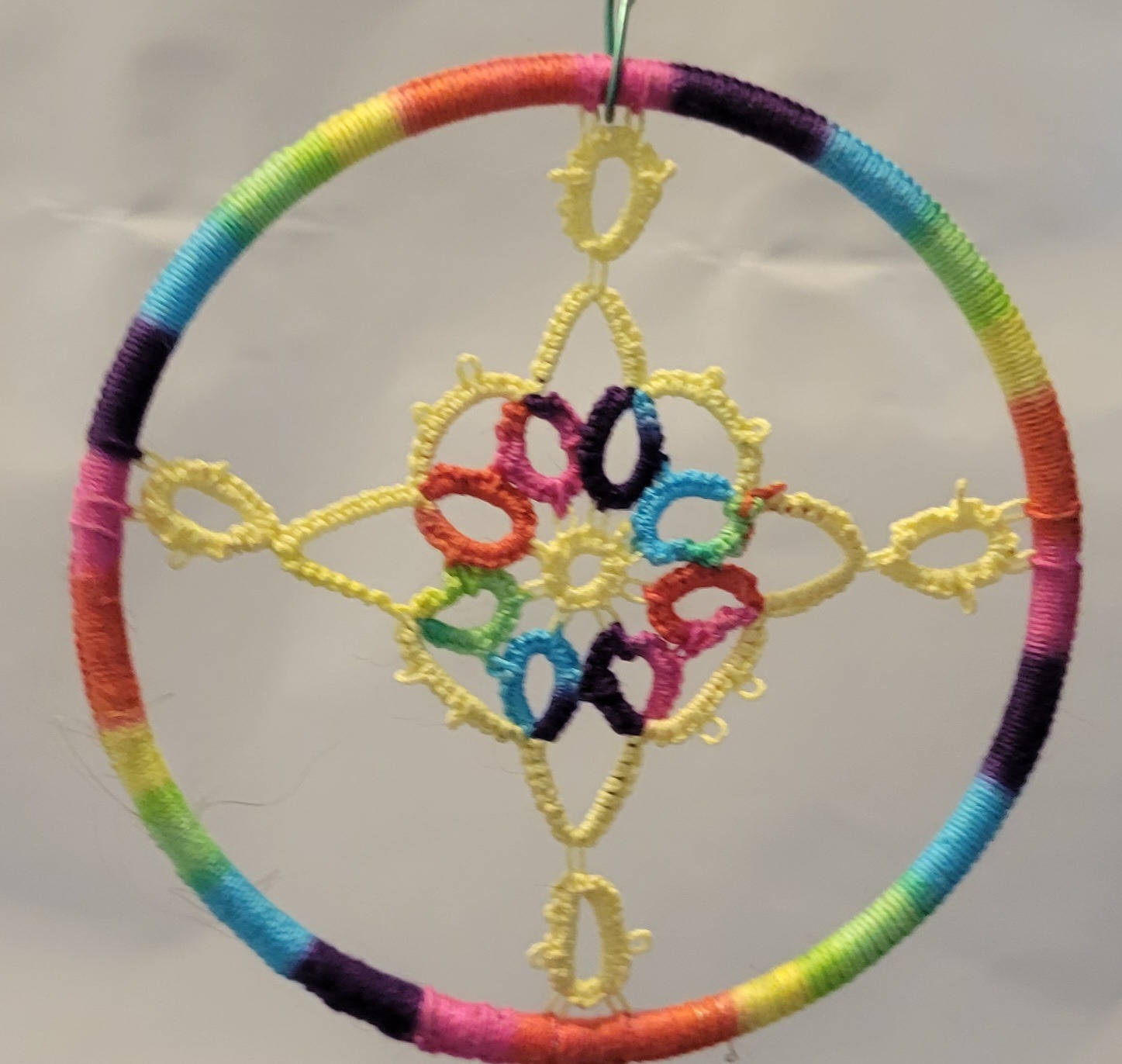 Hand-tatted yellow lace mandala displayed in a rainbow-wrapped circular hoop. The floral-inspired design features delicate symmetrical lacework with colorful accents and measures approximately 3.2 inches in diameter.
