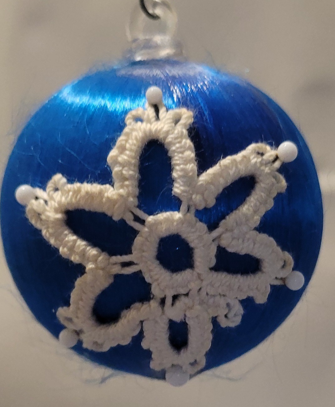 Blue satin ornament decorated with an white hand-tatted snowflake lace motif accented with white pins.