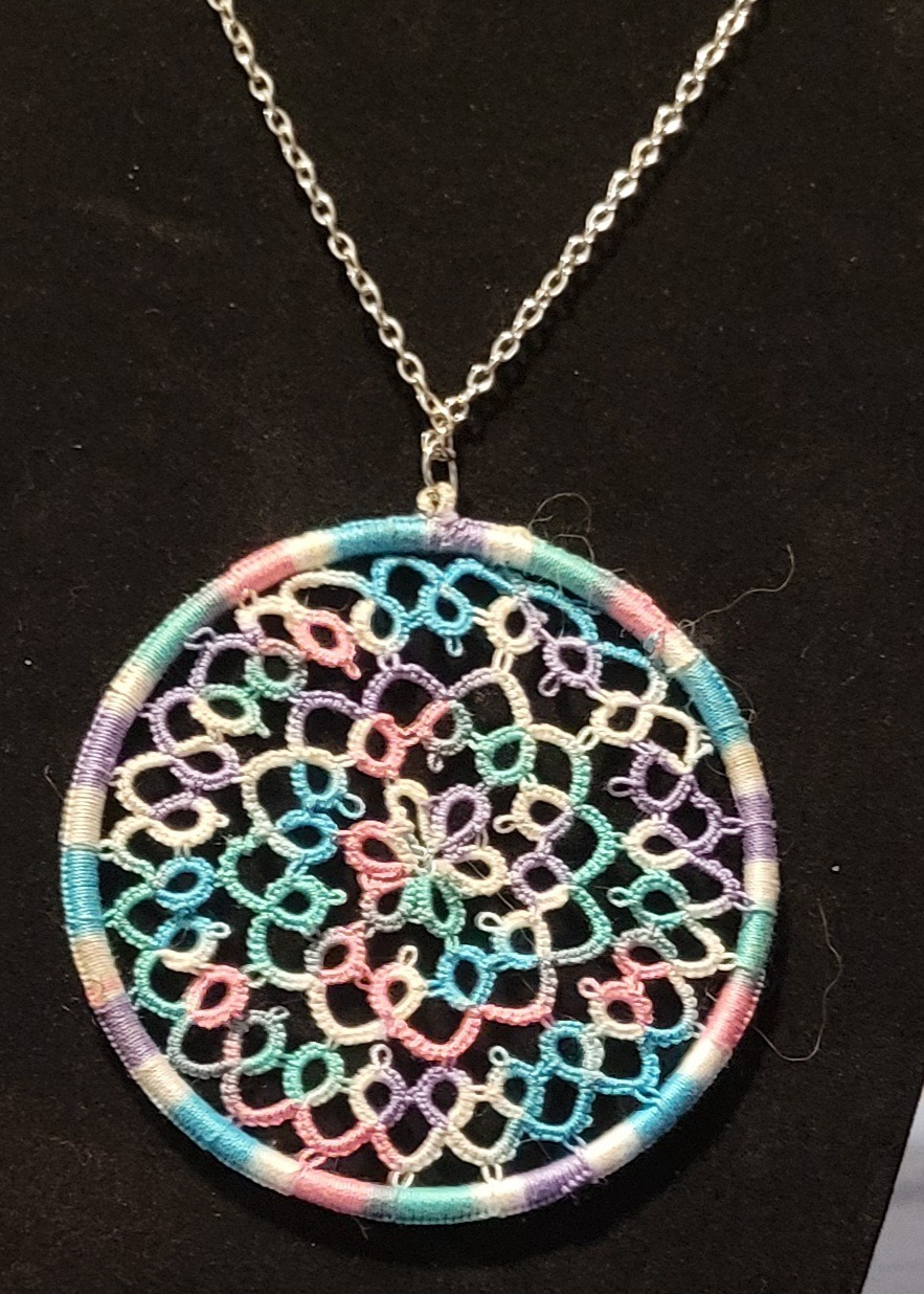 Round pastel hand-tatted lace medallion pendant in aqua, lavender, pink, and white on a 20-inch silver-tone chain displayed against a black background.
