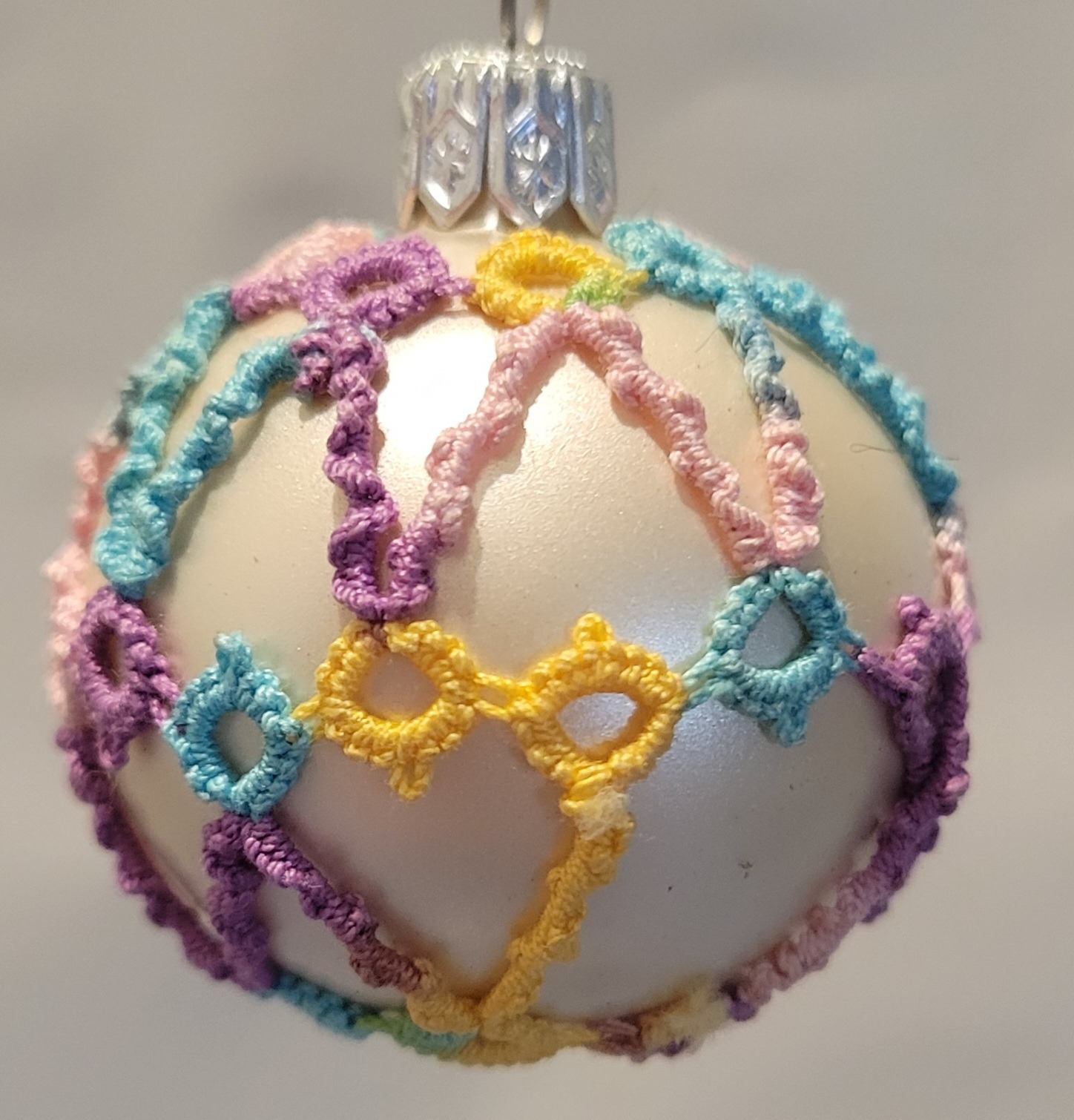 Small pearl-white ornament covered with pastel rainbow hand-tatted lace in delicate looping patterns.