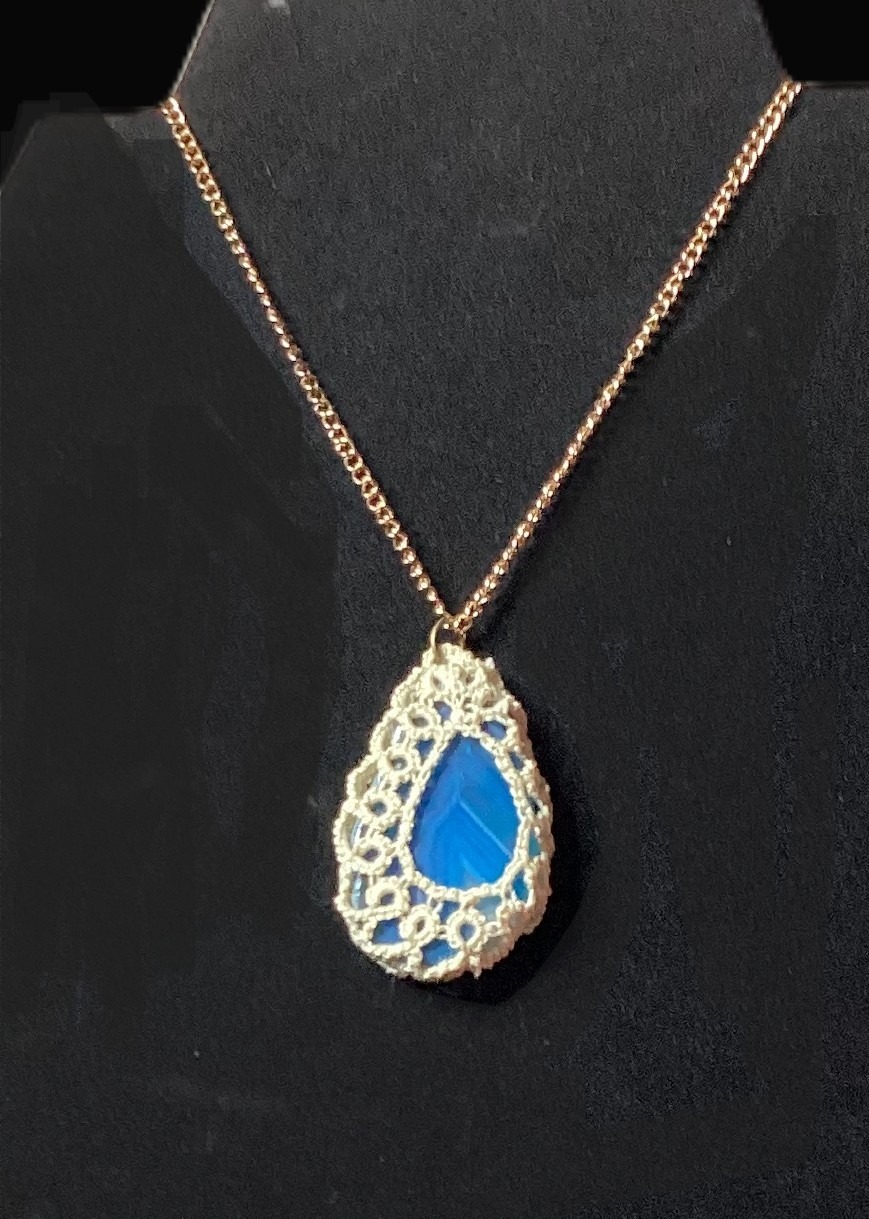Blue agate pendant wrapped in ivory hand-tatted lace on a 20-inch chain displayed against a black jewelry bust.