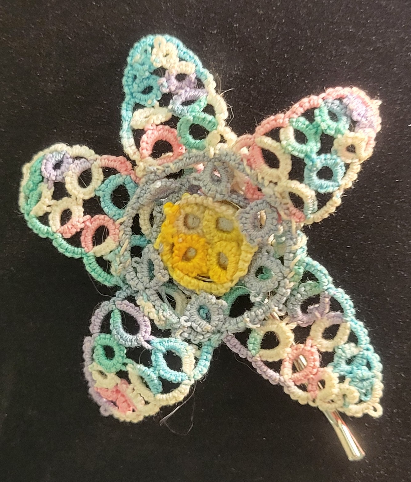 Hand-tatted floral hair pin with pastel lace petals in aqua, pink, lavender, and cream surrounding a yellow flower center, mounted on a metal hair pin, 3 inches in diameter.