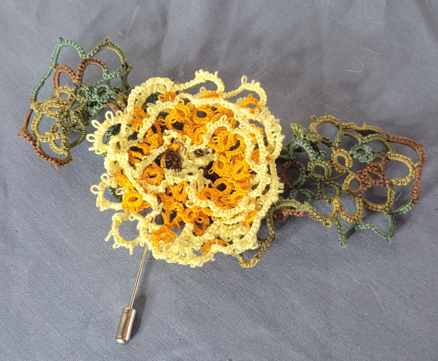 Hand-tatted golden flower hat and lapel pin with cream petals and variegated green leaves on a silver pin back.