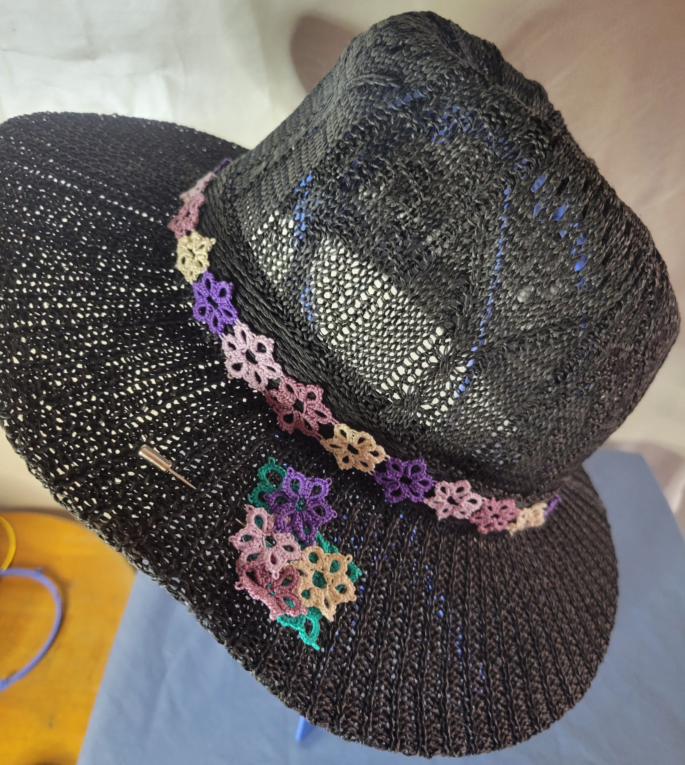 Black woven hat decorated with a hand-tatted wildflower lace hat band and matching floral bouquet hat pin in shades of purple, pink, cream, and green.